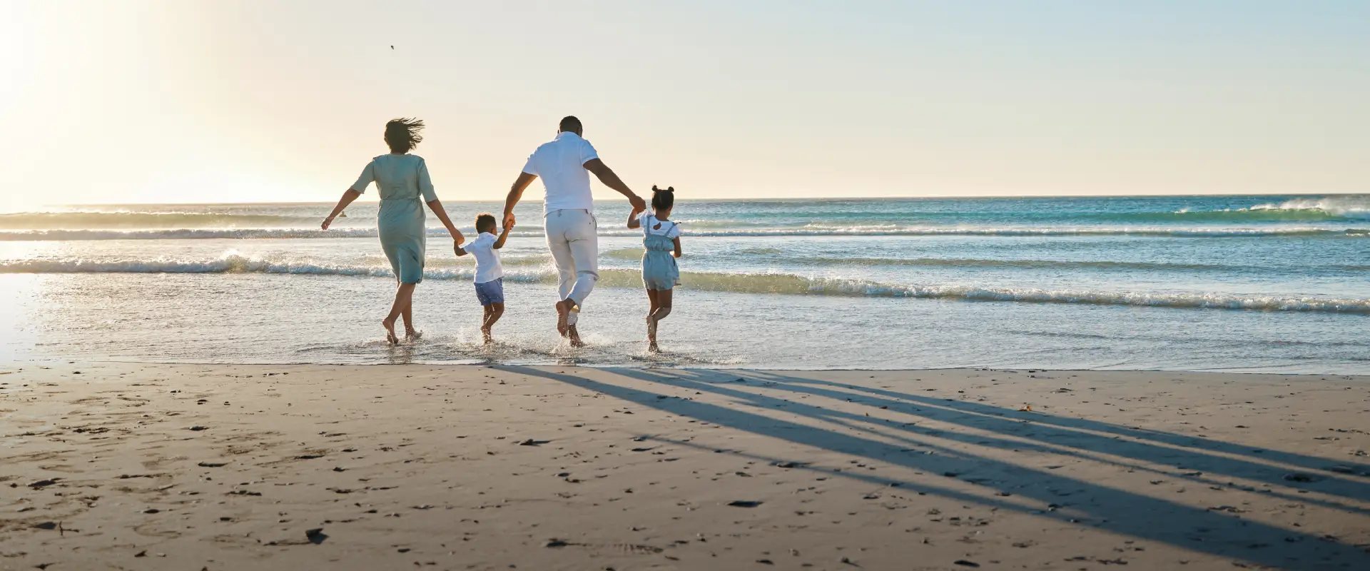 A Black family of four, parents and two children, walk hand-in-hand across a sunlit beach, their long shadows trailing behind them.
